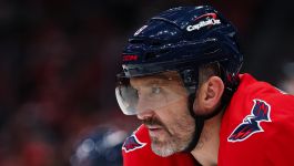 Na zdjęciu widać zawodnika Washington Capitals, Aleksandra Owieczkina, podczas meczu ligi NHL. Ubrany jest w charakterystyczny czerwony strój drużyny, z wyraźnie widocznym logo Capitals na ramieniu i kasku. Owieczkin wygląda na skupionego, być może przygotowuje się do wejścia na lód lub podczas przerwy między zmianami. Wyraz twarzy sugeruje koncentrację i zaangażowanie w grę – być może myśli o kolejnej okazji do zdobycia historycznej, 900. bramki.