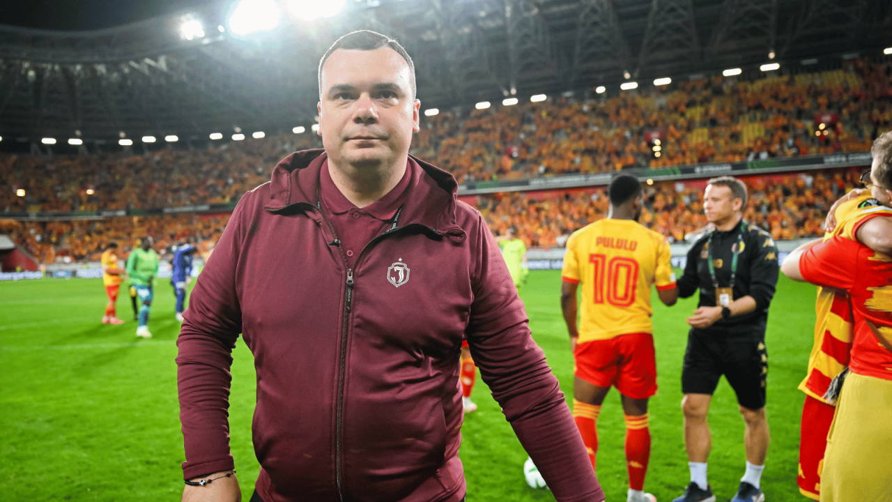 Trener Jagiellonii Białystok Adrian Siemieniec stoi na murawie stadionu, prawdopodobnie po meczu lub podczas rozgrzewki, ubrany w bordową bluzę z kapturem i zamkiem błyskawicznym. Patrzy prosto w kamerę. W tle widoczni są inni piłkarze (jeden w żółto-czerwonej koszulce z numerem 10 i napisem "PULULU") oraz trybuny wypełnione kibicami w żółto-czerwonych barwach (fot. Getty).