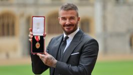 David Beckham z odznaczeniem szlacheckim (fot. Getty Images) Zdjęcie Davida Beckhama w ciemnym garniturze i krawacie, stojącego na zewnątrz na tle historycznego budynku (prawdopodobnie Zamku Windsor). Uśmiecha się i trzyma otwarte, czerwone etui z medalem Orderu Imperium Brytyjskiego (prawdopodobnie Rycerza Kawalera lub Komandora Orderu). Na rękach widać tatuaże. Beckham otrzymał tytuł szlachecki od króla Karola III za wkład w sport i działalność charytatywną.