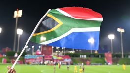 Flaga Republiki Południowej Afryki (fot. Getty Images) Na pierwszym planie widzimy rękę unoszącą flagę Republiki Południowej Afryki, dumnie powiewającą na tle rozświetlonego stadionu. Nocna sceneria, intensywne światła reflektorów i zawodnicy w żółtych strojach na murawie sugerują atmosferę międzynarodowego meczu piłkarskiego. W tle dostrzec można tłum kibiców oraz kolorowe banery, które dodają energii i podkreślają sportowe emocje. Zdjęcie emanuje narodową dumą i sportowym entuzjazmem (fot. Getty Images)