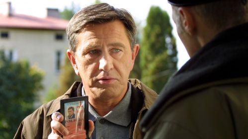 Mężczyzna w średnim wieku stoi na zewnątrz, w słoneczny dzień, i pokazuje legitymację policyjną rozmówcy. Jego wyraz twarzy jest skupiony i stanowczy, co podkreśla jego zawodowy autorytet. To Andrzej Gajewski, grany przez Jerzego Radziwiłowicza w serialu „Glina”, w scenie, która oddaje charakterystyczny dla tej produkcji realizm i surowość policyjnej codzienności.